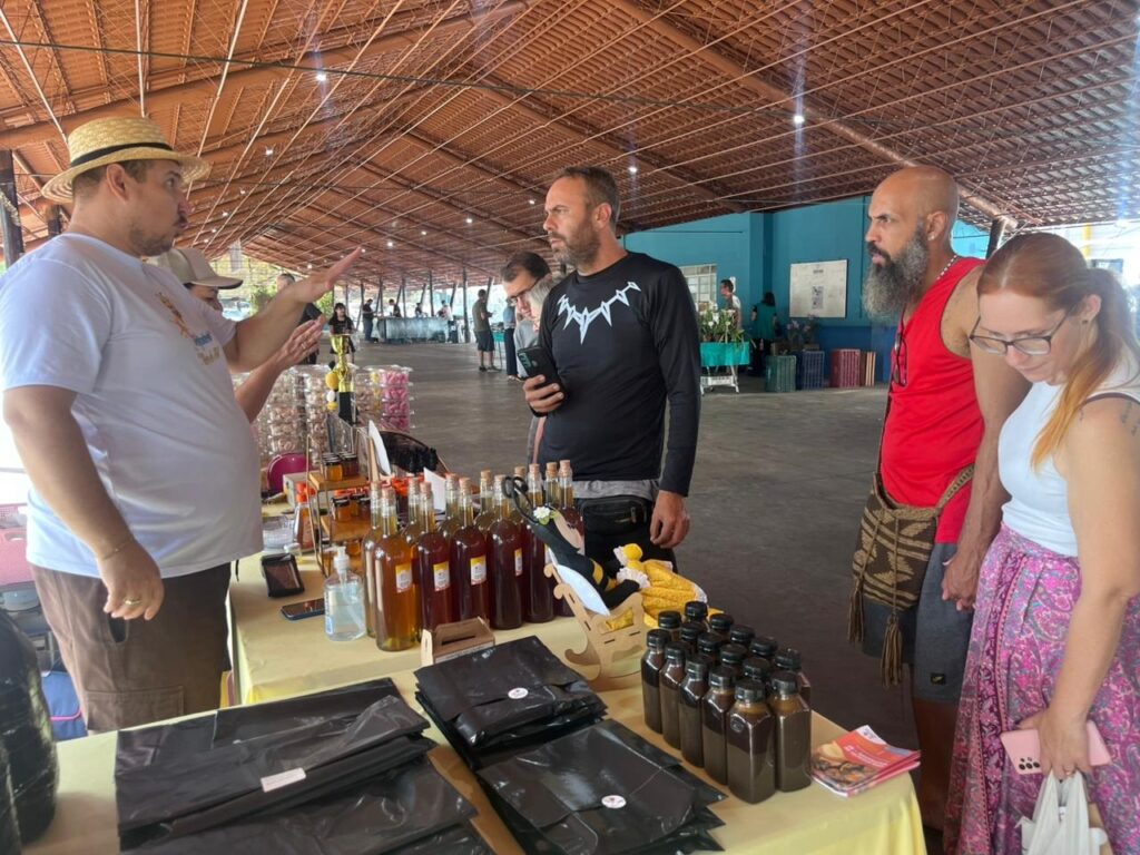A imagem mostra uma cena animada em um mercado ou feira coberta, provavelmente no Brasil, dado o estilo e os produtos. No centro da foto, há um vendedor usando um chapéu de palha e uma camiseta branca, que está gesticulando e conversando com três clientes do outro lado da mesa. Na mesa, há várias garrafas de vidro contendo líquidos de cores variadas, que parecem ser mel ou xaropes artesanais. Também há potes menores e embalagens pretas, além de um pequeno expositor de madeira. Os clientes parecem interessados: um homem de camiseta preta com estampa branca, um homem de regata vermelha com barba grisalha e uma mulher de cabelos ruivos com óculos e saia florida. O ambiente é amplo, com uma estrutura metálica e teto de telhas marrons, permitindo a entrada de bastante luz natural. Ao fundo, é possível ver outras barracas e pessoas, reforçando a ideia de uma feira ou evento comunitário. A atmosfera é de interação social e comércio local, com um clima descontraído e acolhedor.