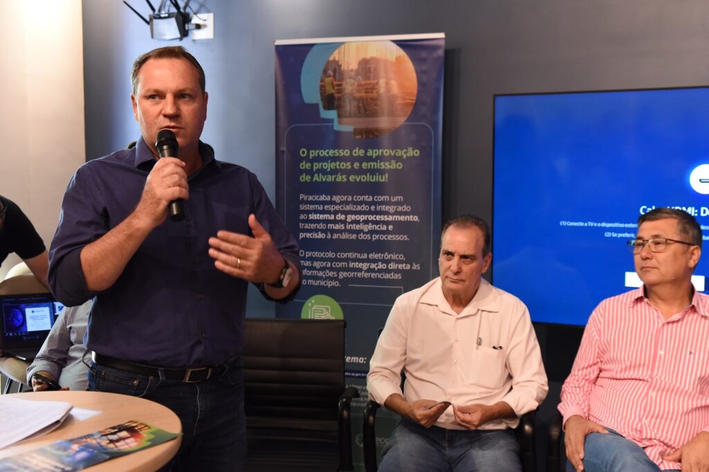 A imagem mostra um homem falando ao microfone durante um evento ou apresentação. Ele está vestido com uma camisa azul escura e parece estar explicando algo, gesticulando com a mão direita. Ao seu lado, sentados, há dois homens, um com camisa branca e outro com camisa listrada rosa e branca, que parecem estar ouvindo atentamente. Atrás deles, há um banner vertical com texto em português que fala sobre a modernização do processo de aprovação de projetos e emissão de alvarás em Piracicaba, destacando a utilização de um sistema integrado de geoprocessamento. À direita, há uma tela azul com texto branco, provavelmente usada para apoiar a apresentação. Na mesa à frente, há alguns documentos e um folheto, indicando que se trata de um ambiente formal, possivelmente uma coletiva de imprensa ou uma reunião oficial. A iluminação é clara, típica de ambientes internos corporativos.