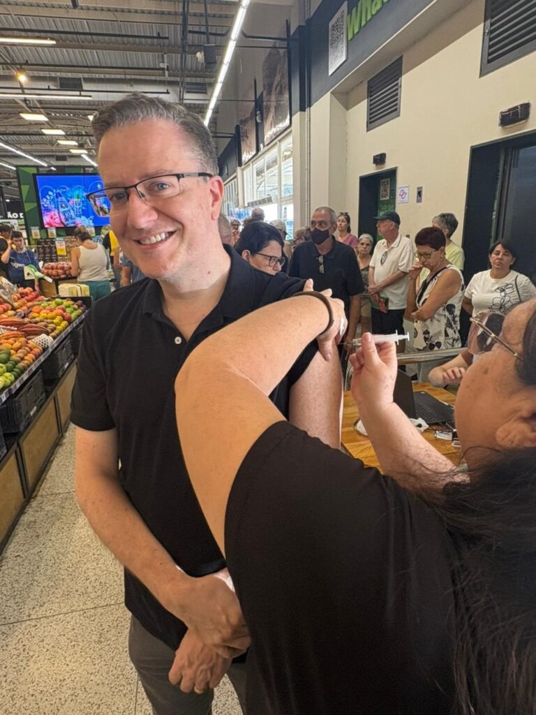 A imagem mostra um homem sorridente recebendo uma vacina no braço, em um ambiente que parece ser um supermercado. Ele usa óculos e uma camisa preta, e está olhando diretamente para a câmera. Ao seu lado, uma pessoa está aplicando a vacina com uma seringa. Ao fundo, há prateleiras com frutas e verduras e outras pessoas, algumas usando máscaras, aguardando na fila. O ambiente é iluminado artificialmente, típico de um espaço interno de comércio. A cena transmite a ideia de uma ação de vacinação sendo realizada de forma acessível e comunitária, dentro de um local de compras cotidiano.