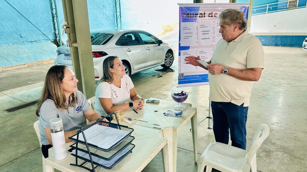 A imagem mostra um evento de recrutamento de emprego realizado em um espaço interno que parece ser uma garagem ou estacionamento coberto, com paredes pintadas de azul claro e piso de concreto. No centro da cena, há uma mesa branca de plástico com três pessoas interagindo: duas mulheres sentadas e um homem mais velho em pé, conversando com elas. O homem usa uma camisa polo bege e calça jeans, e parece estar explicando algo ou fazendo uma pergunta, segurando um pequeno papel. Uma das mulheres está vestida com uma camiseta cinza com o logo "S2B" e sorri para ele, enquanto a outra, de camiseta branca, observa atentamente. Sobre a mesa, há alguns objetos, como uma bandeja com papéis, um copo térmico branco, um recipiente com petiscos roxos e um celular. Ao fundo, há um banner com o título "Mural de Vagas", que lista algumas vagas de emprego disponíveis, como açougueiro, balconista de açougue, designer gráfico, marketing e técnico em segurança do trabalho. Também é possível ver um carro prata estacionado atrás do grupo. A iluminação é natural, indicando que o evento ocorre durante o dia. O ambiente transmite uma atmosfera profissional, porém informal, típica de um processo seletivo comunitário ou local.