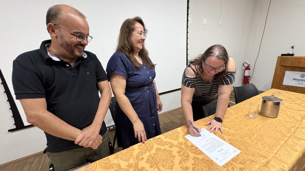 A imagem mostra uma cerimônia formal de posse ou assinatura de documento, realizada em um ambiente interno. Há três pessoas em destaque: um homem à esquerda, usando óculos, barba, camiseta preta e calça cáqui, que está de pé com as mãos juntas, sorrindo. No centro, uma mulher de cabelos longos e castanhos, usando óculos e vestido azul escuro, também sorrindo. À direita, uma mulher com óculos, camiseta listrada preta e branca, está sentada ou inclinada sobre uma mesa com toalha dourada, assinando um documento com uma caneta azul. Ela tem uma tatuagem visível no ombro direito e usa um relógio preto. Ao fundo, há um telão branco, um púlpito de madeira com o brasão e o nome "Piracicaba", um extintor de incêndio vermelho e paredes brancas. Sobre a mesa, além do documento, há um copo com água e um balde de gelo metálico. A atmosfera parece formal e positiva, indicando um evento oficial, provavelmente uma cerimônia de posse, como sugere o texto parcialmente visível "TERMO DE POSSE".