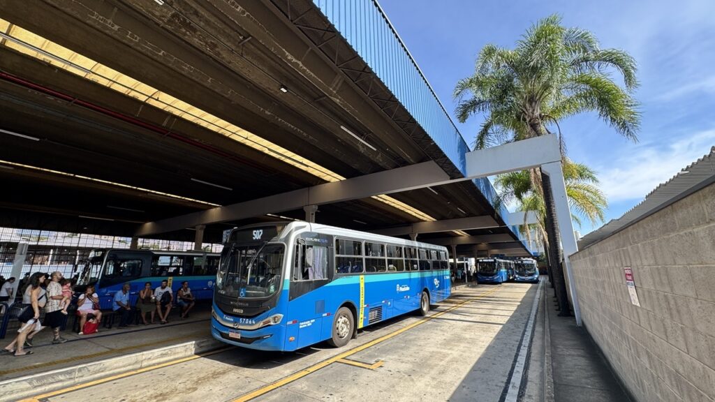 A imagem mostra um terminal de ônibus em um dia ensolarado, com céu azul e poucas nuvens. Em destaque, há um ônibus azul e branco da marca IVECO, identificado pelo número 57064, com a palavra "Piracicaba" escrita na lateral. O ônibus está parado em uma plataforma de embarque sob uma grande cobertura de concreto e metal. Ao lado, outro ônibus semelhante está estacionado, e várias pessoas estão sentadas em bancos ou em pé, aguardando o transporte. Elas vestem roupas leves, adequadas para o clima quente, como camisetas, shorts e sandálias. À direita, há uma parede de tijolos claros e uma palmeira alta. O chão é de concreto com faixas amarelas que indicam os espaços para os ônibus. Ao fundo, outros ônibus estão alinhados. A estrutura do terminal, com suas linhas retas e a cobertura, cria uma sensação de profundidade na imagem. A iluminação é forte, com sombras bem definidas, indicando que a foto foi tirada durante o dia.