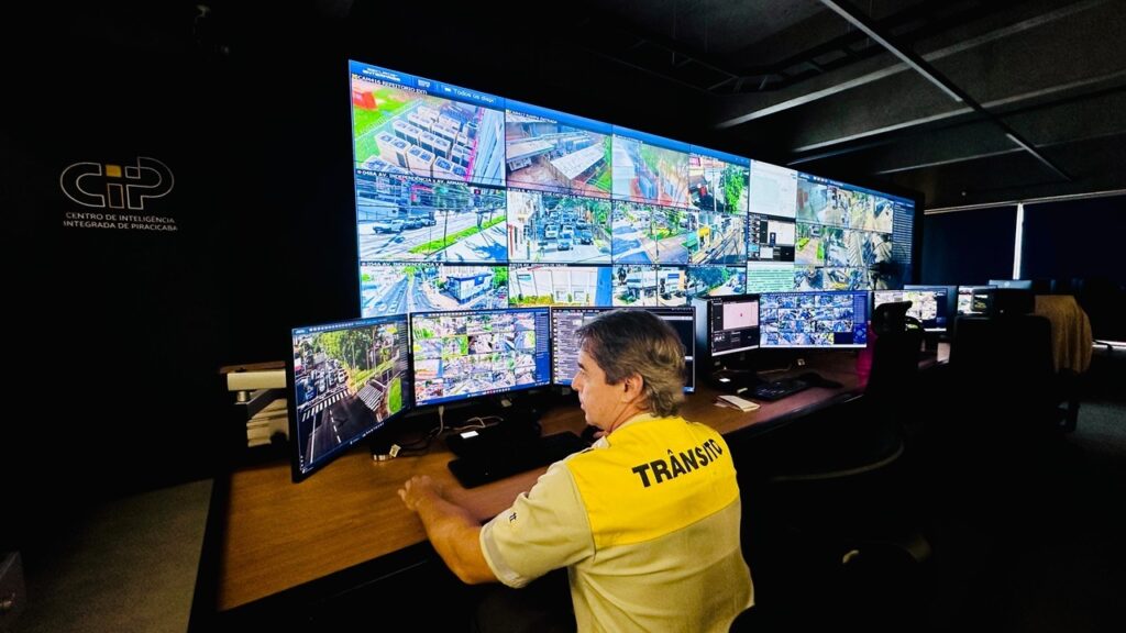 A imagem mostra uma sala de controle moderna, provavelmente dedicada ao monitoramento de trânsito ou segurança pública na cidade de Piracicaba, conforme indicado pelo logo "CIP - Centro de Inteligência Integrada de Piracicaba" na parede preta à esquerda. Na parede principal, há um grande painel de vídeo composto por múltiplas telas que exibem imagens ao vivo de várias câmeras espalhadas pela cidade, mostrando ruas, cruzamentos e áreas externas de edifícios. Em primeiro plano, um operador está sentado de perfil, vestindo uma camisa clara com uma faixa amarela nas costas onde está escrito "TRÂNSITO". Ele está focado em vários monitores de computador posicionados à sua frente, que também exibem imagens de câmeras e mapas, sugerindo que ele está monitorando o tráfego em tempo real. O ambiente é pouco iluminado, com a principal fonte de luz vindo das telas, criando um contraste que destaca a tecnologia e a operação em andamento. A perspectiva da foto é ampla, mostrando tanto o operador quanto a estrutura tecnológica do centro de controle, evidenciando a complexidade e a organização da operação de monitoramento.