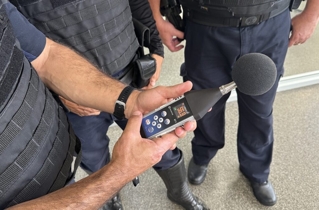 A imagem mostra um close das mãos de uma pessoa segurando um medidor de nível sonoro da marca SVANTEK, modelo 971. O visor do aparelho indica uma leitura de 56,9 decibéis (dB). O medidor está equipado com uma espuma protetora preta na parte superior, que cobre o microfone. Ao redor da pessoa que segura o aparelho, há outras pessoas vestindo uniformes táticos, provavelmente policiais ou agentes de segurança, com coletes e cintos com equipamentos. O ambiente parece ser externo, com o chão de concreto e uma parede de vidro ao fundo. A cena sugere uma inspeção ou medição do nível de ruído realizada por autoridades.
