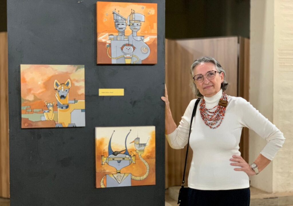 A imagem mostra uma mulher em uma galeria de arte, posicionada ao lado de uma parede preta onde estão expostas três obras de arte quadradas, dispostas verticalmente. Ela está sorrindo, usando óculos, uma blusa branca de gola alta e uma saia ou calça preta. Também usa um colar grande e colorido, em tons de vermelho e branco. As três obras têm um estilo ilustrativo com cores predominantemente laranja, amarelo e cinza, e parecem representar figuras futuristas ou robóticas. A pintura superior mostra duas figuras robóticas e uma figura humana menor; a do meio à esquerda retrata um cão robótico; e a inferior apresenta um personagem robótico com antenas longas e uma criatura semelhante a um pássaro. Ao lado da mulher, há uma pequena etiqueta amarela, e no canto inferior esquerdo da imagem está escrito “Regina Silveira - Brazil”, indicando provavelmente o nome da artista ou a origem das obras. O ambiente é interno, bem iluminado, com paredes claras e divisórias de madeira.