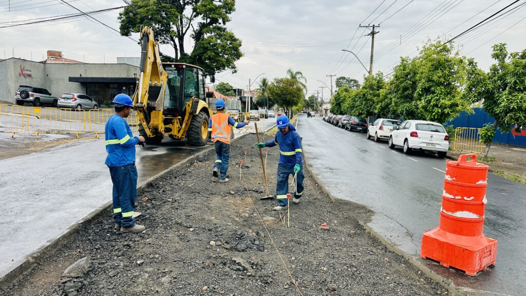 A imagem mostra uma obra de construção em uma via urbana durante um dia nublado. Três trabalhadores estão vestidos com uniformes de segurança azul e laranja e capacetes, trabalhando em uma faixa estreita de terreno escavado, onde há uma mistura de cascalho e terra. Um deles está próximo a uma retroescavadeira amarela, que está posicionada ao lado da faixa de obra. Dois outros trabalhadores estão mais adiante, um segurando uma ferramenta e o outro caminhando. À esquerda da faixa de obra, há uma pista de asfalto que está bloqueada por barreiras metálicas amarelas, indicando que o trânsito está impedido naquele trecho. À direita, há uma pista pavimentada com carros estacionados, incluindo um Volkswagen Gol branco com a placa OAA 9328. Também há um cone de trânsito laranja próximo à faixa de construção. Ao fundo, é possível ver postes de energia, árvores verdes e construções residenciais ou comerciais, além de uma placa amarela com o nome "XAVIER CAR CENTER". O cenário sugere que a obra está acontecendo em uma área urbana, possivelmente para manutenção ou instalação de infraestrutura, como rede de água, esgoto ou cabeamento.