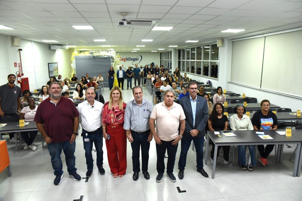 A imagem mostra uma sala de aula ou auditório moderno e bem iluminado, com várias pessoas presentes. No primeiro plano, há seis adultos em pé, alinhados e voltados para a câmera, parecendo ser os organizadores ou palestrantes do evento. Eles estão vestidos de maneira casual e profissional. Atrás deles, há várias fileiras de pessoas sentadas em mesas longas, provavelmente participantes de uma palestra, curso ou reunião. A sala possui um teto com painéis de iluminação embutidos e um projetor fixado no centro. Nas paredes, há janelas grandes com persianas brancas que permitem a entrada de luz natural. À esquerda, há uma porta aberta e um extintor de incêndio preso à parede. No fundo, pode-se ver um painel com o logotipo "Kingklinger" e as palavras "NEW YORK 1990". O ambiente transmite uma atmosfera profissional e organizada, típica de treinamentos corporativos, workshops ou eventos educacionais.