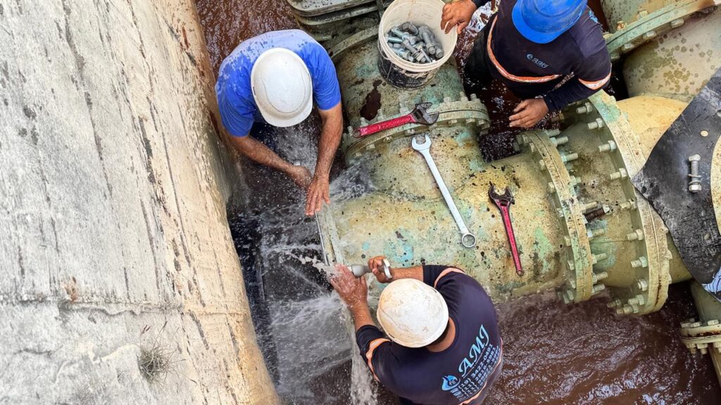 A imagem mostra três trabalhadores de manutenção realizando reparos em um grande cano de água pressurizada dentro de uma vala de concreto. O cano é de metal pintado de verde claro, com juntas flangeadas aparafusadas. De uma das juntas à esquerda, há um vazamento forte de água, que está sendo contido ou controlado por um dos trabalhadores que veste camiseta azul e capacete branco. Outro trabalhador, na parte inferior da imagem, com camiseta preta estampada com o logo "AMJ" e capacete branco, está utilizando uma ferramenta para apertar uma conexão, enquanto é atingido pelo jato de água. Um terceiro trabalhador, no canto superior direito, também com camiseta preta e capacete azul, está estendendo o braço em direção ao cano, possivelmente ajudando no reparo. Sobre o cano estão várias ferramentas, incluindo duas chaves de grifo vermelhas e uma chave inglesa prateada. Próximo ao topo da imagem há um balde branco contendo vários parafusos grandes. A vala contém água suja e lamacenta, indicando que o reparo está ocorrendo em condições difíceis. Os trabalhadores parecem estar em uma situação de manutenção urgente, lidando com um vazamento significativo em um ambiente industrial. O logo "AMJ" e números de telefone estão visíveis nas camisetas dos trabalhadores, sugerindo o nome da empresa responsável pelo serviço.