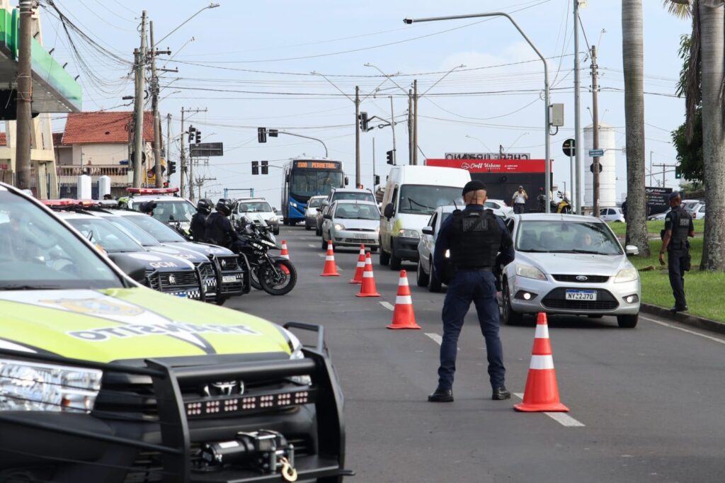 A imagem mostra uma operação de fiscalização de trânsito em uma via urbana movimentada. No centro da cena, um agente de segurança, vestindo uniforme escuro e colete tático, está posicionado de costas, orientando o fluxo de veículos. A pista está parcialmente bloqueada por cones laranja, organizados em linha para canalizar os carros. À direita, um veículo branco aguarda abordagem, enquanto outros carros formam uma fila. Ao fundo, é possível ver um ônibus e mais veículos transitando lentamente.