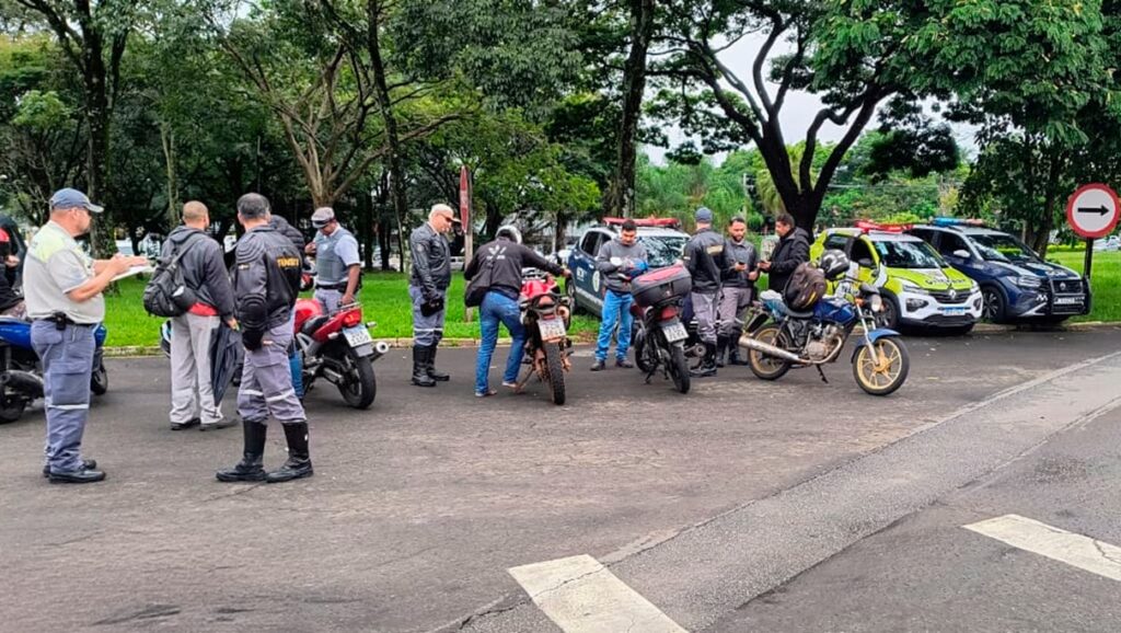 A imagem mostra uma operação de fiscalização de trânsito ao ar livre, provavelmente em um parque ou área verde, com vários policiais e agentes de trânsito interagindo com motociclistas. Há ao menos três veículos policiais estacionados, incluindo um Renault Duster branco com detalhes em amarelo e um SUV azul escuro, ambos com sirenes no teto. Na cena, alguns motociclistas estão parados ao lado de suas motos, enquanto os agentes, vestidos com uniformes que indicam "TRANSITO" e "GCM" (Guarda Civil Municipal), realizam a abordagem e anotam informações, possivelmente conferindo documentos ou aplicando multas. Um policial está de costas, usando uma jaqueta preta com a palavra "TRANSITO" em amarelo. Outro agente está escrevendo em uma prancheta. O ambiente é cercado por árvores e vegetação, com um caminho pavimentado onde ocorre a operação. No canto direito, há uma placa de trânsito circular vermelha com uma seta preta apontando para a direita, indicando uma direção obrigatória ou restrição de circulação. A atmosfera geral da imagem é de controle e fiscalização do trânsito, com uma ação organizada e coordenada dos agentes para garantir a segurança nas vias.
