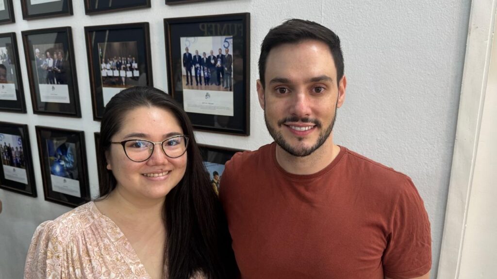 A imagem mostra um homem e uma mulher lado a lado, sorrindo para a câmera. Eles estão em um ambiente interno, com uma parede branca ao fundo decorada com várias fotografias emolduradas. A mulher, à esquerda, tem cabelos longos e escuros, usa óculos e uma blusa clara com estampa. O homem, à direita, tem cabelo curto, barba e está vestido com uma camiseta de cor ferrugem. O cenário sugere um ambiente profissional ou acadêmico, possivelmente um escritório ou corredor, devido às fotos emolduradas que parecem comemorar marcos ou eventos importantes. A iluminação é natural e o clima geral da foto é amigável e acolhedor.