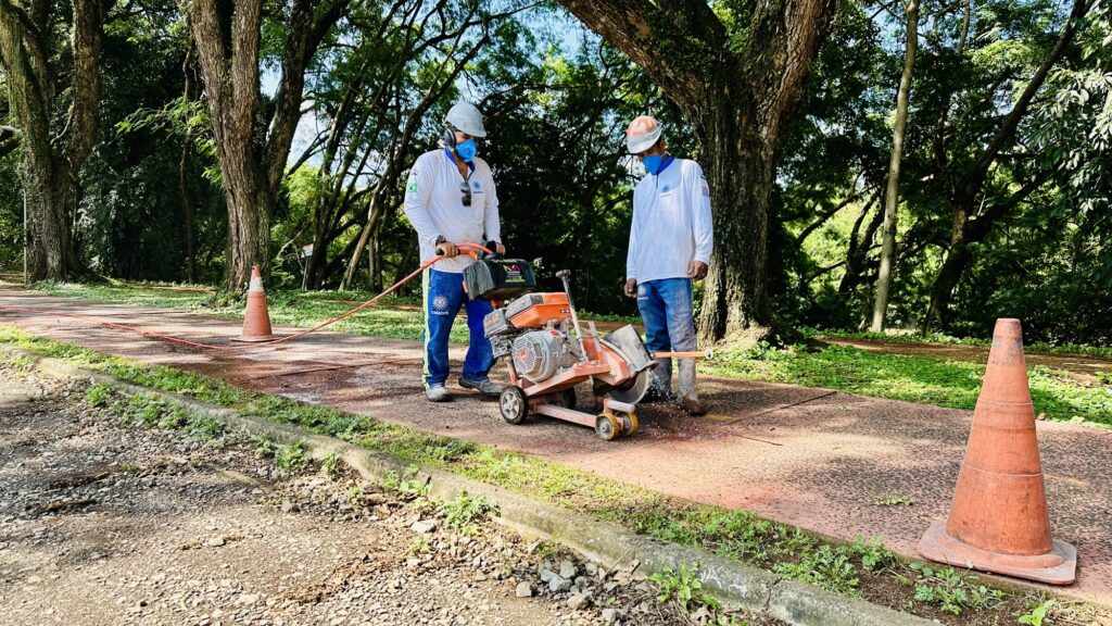 A imagem mostra dois trabalhadores da construção civil em um ambiente ao ar livre, provavelmente em um parque ou área arborizada, realizando a manutenção de uma pista pavimentada. Ambos vestem uniformes brancos com a logo "celebre" estampada, capacetes brancos, máscaras azuis e protetores auriculares. Um dos trabalhadores está operando uma serra de piso, que parece ser uma serra de corte para concreto, enquanto o outro está próximo, observando ou auxiliando. A serra é de cor laranja e está posicionada sobre a pista, que é delimitada por cones laranja, indicando uma área de trabalho. Ao fundo e ao redor, há muitas árvores, e a luz do sol passa pelas folhas, criando um efeito de luz e sombra no chão. A imagem transmite uma sensação de cuidado e profissionalismo durante a realização de um serviço de manutenção em uma área pública.