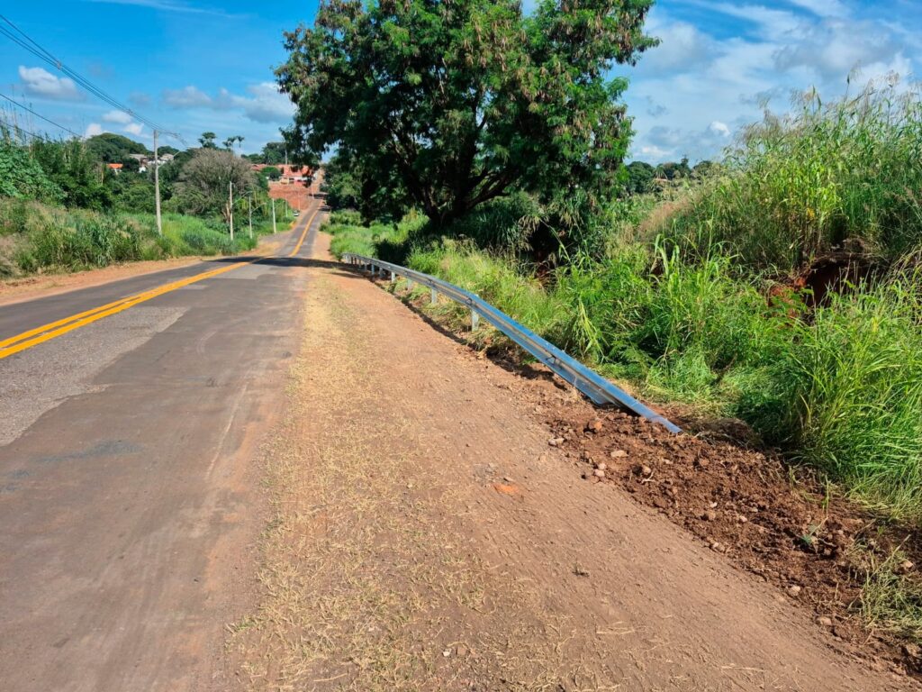 A imagem mostra uma estrada asfaltada em área rural, com duas faixas demarcadas por linhas amarelas no centro. À direita da estrada, há um guarda-corpo metálico danificado, que parece estar afundando ou mal fixado no chão de terra ao lado da pista. O entorno é bastante verde, com vegetação densa e arbustos ao longo da estrada. À esquerda, postes de energia acompanham o trajeto da via. No fundo, é possível ver algumas casas com telhados laranjas, situadas em uma elevação. O céu está azul com algumas nuvens brancas, indicando um dia ensolarado. A imagem transmite uma sensação de tranquilidade típica do campo, embora o guarda-corpo danificado sugira necessidade de manutenção na estrada.