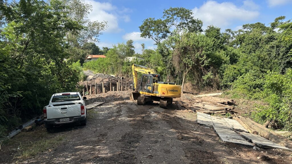 A imagem mostra um cenário de construção em uma área rural, sob um céu azul com algumas nuvens. No centro da imagem, há uma escavadeira amarela da marca LiuGong, modelo 915E, que parece estar trabalhando na construção ou reparo de uma ponte ou estrada de terra. À esquerda, há uma caminhonete branca Chevrolet S10 estacionada, com placa EQB-3448. Ao redor, é possível ver troncos de madeira, postes verticais de madeira e blocos de concreto que provavelmente fazem parte da obra. Ao fundo, há muitas árvores verdes e uma casa simples, reforçando o ambiente rural. A cena transmite uma atividade de infraestrutura em andamento em uma região mais afastada.