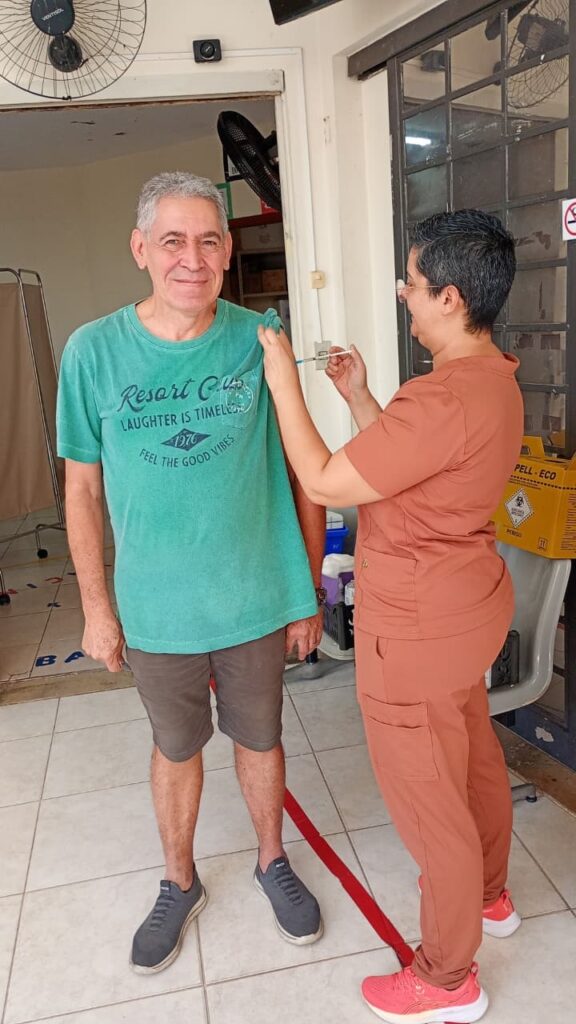A imagem mostra um homem idoso recebendo uma vacina no braço esquerdo, aplicada por uma profissional de saúde. O homem está sorrindo e usa uma camiseta verde-azulada com a frase "Resort Club LAUGHTER IS TIMELESS 1976 FEEL THE GOOD VIBES" e shorts cinza. A profissional está vestida com um uniforme cor de terracota e tênis rosa, concentrada na aplicação da vacina. Eles estão em um ambiente interno, provavelmente uma clínica ou posto de saúde, com piso branco e paredes claras. Ao lado, há um recipiente amarelo para descarte de materiais perfurocortantes com a marca "PELL-ECO" e a palavra "PERIGO". Na parede, um ventilador da marca "VENTISOL" está instalado. A cena transmite uma sensação de cuidado e segurança, relacionada a uma campanha de vacinação.
