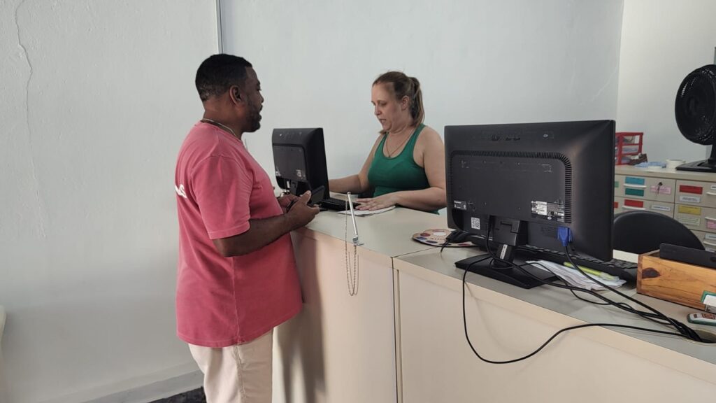 A imagem mostra um ambiente de escritório ou recepção, onde um homem e uma mulher estão interagindo. O homem, que veste uma camiseta rosa e calça cáqui, está em pé do lado de fora do balcão, segurando um celular. A mulher, com cabelo loiro preso em um rabo de cavalo e usando uma regata verde, está sentada atrás do balcão, olhando para baixo, possivelmente em um computador ou documento. No balcão há dois monitores de computador, teclados, um porta-canetas prateado com uma corrente, uma caixa de madeira e um ventilador preto. Ao fundo, há um armário de arquivos com etiquetas coloridas. A iluminação é clara e o ambiente parece organizado e profissional. A interação sugere um atendimento ou um processo administrativo em andamento.
