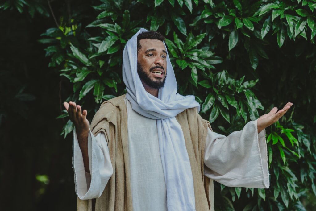 A imagem mostra um homem de pele escura, com barba e cabelo curto e encaracolado, vestido com roupas que lembram trajes históricos ou religiosos do Oriente Médio antigo. Ele usa um manto bege claro e uma faixa branca na cabeça, parecida com um turbante ou keffiyeh. O homem está com os braços abertos, numa postura que pode transmitir um gesto de prece, explicação ou acolhimento. O fundo é composto por uma vegetação densa e verde, sugerindo que ele está ao ar livre, possivelmente em um ambiente natural. A expressão facial dele parece intensa, olhando levemente para cima e para o lado, o que reforça a ideia de um momento de emoção ou comunicação profunda. A foto tem um foco nítido no homem, enquanto o fundo está um pouco desfocado, destacando-o como o centro da imagem.