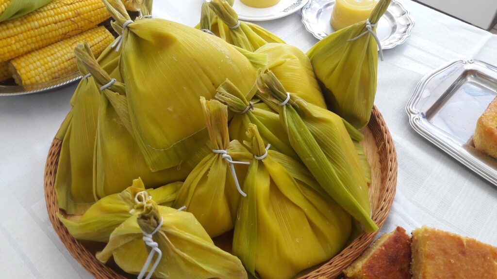 A imagem mostra uma mesa com uma variedade de pratos tradicionais feitos de milho, típicos da culinária brasileira, possivelmente relacionados a uma festa junina. No centro, há uma grande bandeja de palha contendo várias pamonhas, que são envoltas em folhas de milho e amarradas com barbante branco. Ao redor da bandeja, há pratos com outras comidas à base de milho: no canto superior esquerdo, espigas de milho cozidas; no canto superior direito, pequenas porções de curau, um doce cremoso de milho; e no canto inferior direito, pedaços de bolo de milho dourado. A mesa está coberta por uma toalha branca, e a iluminação é clara, destacando as cores vibrantes dos alimentos. A imagem transmite uma sensação de festa e tradição, valorizando a diversidade de pratos feitos com milho.