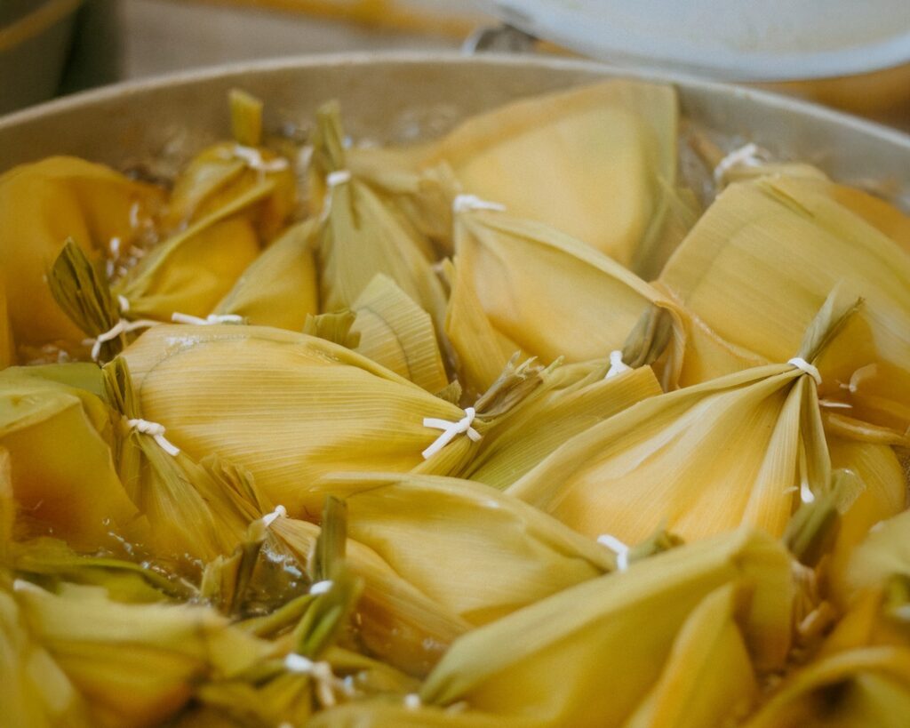 A imagem mostra uma vista aproximada e de cima para baixo de várias pamonhas sendo cozidas em uma panela grande de metal. Cada pamonha está envolto em folhas de milho amarelo-esverdeadas, amarradas firmemente com um cordão branco, formando pequenos pacotes.
