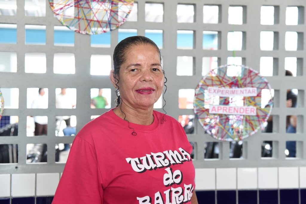 A imagem mostra uma mulher de meia-idade com pele morena e cabelo escuro preso em um rabo de cavalo, com alguns fios cacheados soltos ao redor do rosto. Ela sorri levemente e veste uma camiseta rosa vibrante com a frase "TURMA do BAIRRO" escrita em branco na frente. Ela está posicionada no centro da imagem, em frente a uma parede cinza de concreto que possui uma grade com pequenas aberturas quadradas. Através dessas aberturas, é possível ver um céu azul e algumas pessoas desfocadas ao fundo. À direita da mulher, há um artesanato colorido circular feito de fios entrelaçados, com duas plaquinhas brancas onde se lê "EXPERIMENTAR" e "APRENDER" em letras vermelhas maiúsculas. Outra peça semelhante aparece parcialmente no canto superior esquerdo. A iluminação é clara e uniforme, sugerindo que a foto foi tirada durante o dia, em um local aberto ou bem iluminado. A composição da foto é um retrato simples, com foco nítido na mulher, enquanto o fundo está levemente desfocado.