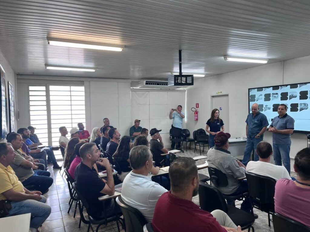 A imagem mostra uma sala de aula ou auditório onde um grupo de pessoas está participando de uma apresentação ou reunião. Três pessoas estão na frente da sala, aparentemente apresentando ou falando para o público. Uma delas está fazendo um gesto com a mão, indicando que está explicando algo. O público, composto por cerca de vinte pessoas, está sentado em cadeiras dispostas em filas, todas voltadas para os apresentadores. A sala é iluminada por luzes fluorescentes no teto, que é branco com painéis. Há uma tela digital grande ao fundo que exibe uma grade de imagens ou gráficos, mas os detalhes não são claramente visíveis. Também é possível ver uma janela com persianas ao lado esquerdo da sala, sugerindo que é dia. Um extintor de incêndio vermelho está fixado na parede ao fundo, próximo a uma porta. As pessoas na plateia vestem roupas casuais, como camisetas e jeans, e parecem atentas à apresentação. Um homem está tirando uma foto ou gravando com um celular. A disposição do ambiente e a postura dos presentes indicam que se trata de um evento profissional, seminário ou oficina educacional.