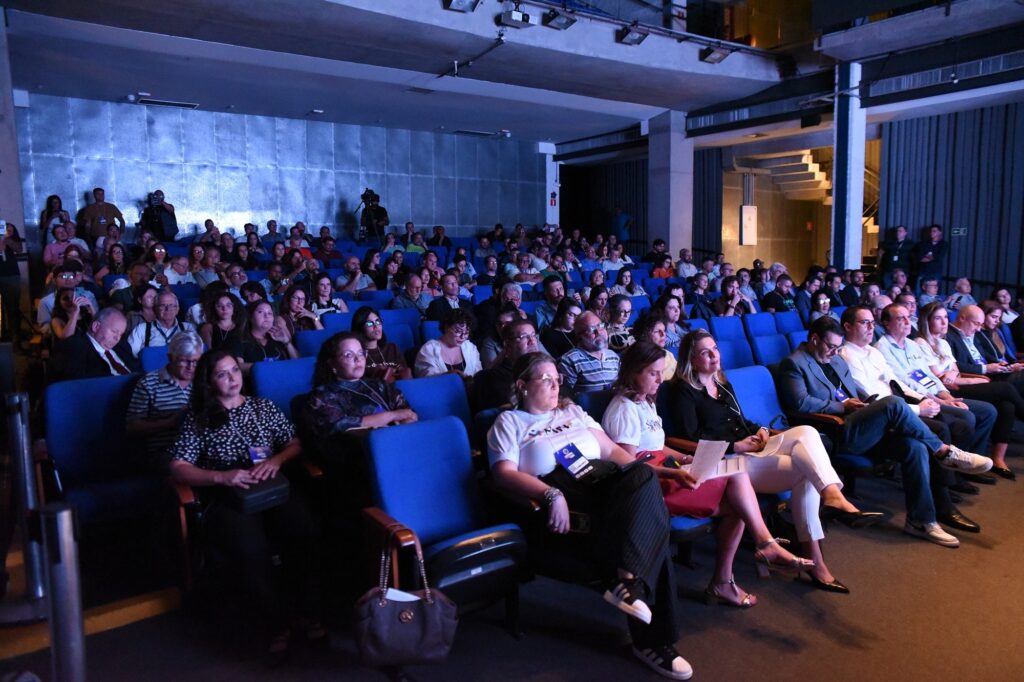 A imagem mostra um auditório cheio de pessoas sentadas em cadeiras estofadas de cor azul, assistindo a um evento formal. A foto foi tirada em um ângulo diagonal, da esquerda para a direita, capturando várias fileiras de espectadores. A maioria está vestida de forma casual ou social, com algumas pessoas usando camisas sociais e outras mais descontraídas. No primeiro plano, uma mulher usa uma camiseta branca com um logo colorido e um crachá pendurado no pescoço. Ao lado dela, homens de terno e camisa clara estão sentados, alguns com postura atenta. O ambiente é iluminado com luz azulada, provavelmente vinda do palco ou de uma tela à frente, fora do campo da câmera. Ao fundo, pode-se ver câmeras em tripés, indicando que o evento está sendo gravado. O teto é de concreto aparente, com luminárias e dutos expostos, conferindo um aspecto moderno e industrial ao espaço. A atmosfera geral é de concentração e interesse por parte do público.