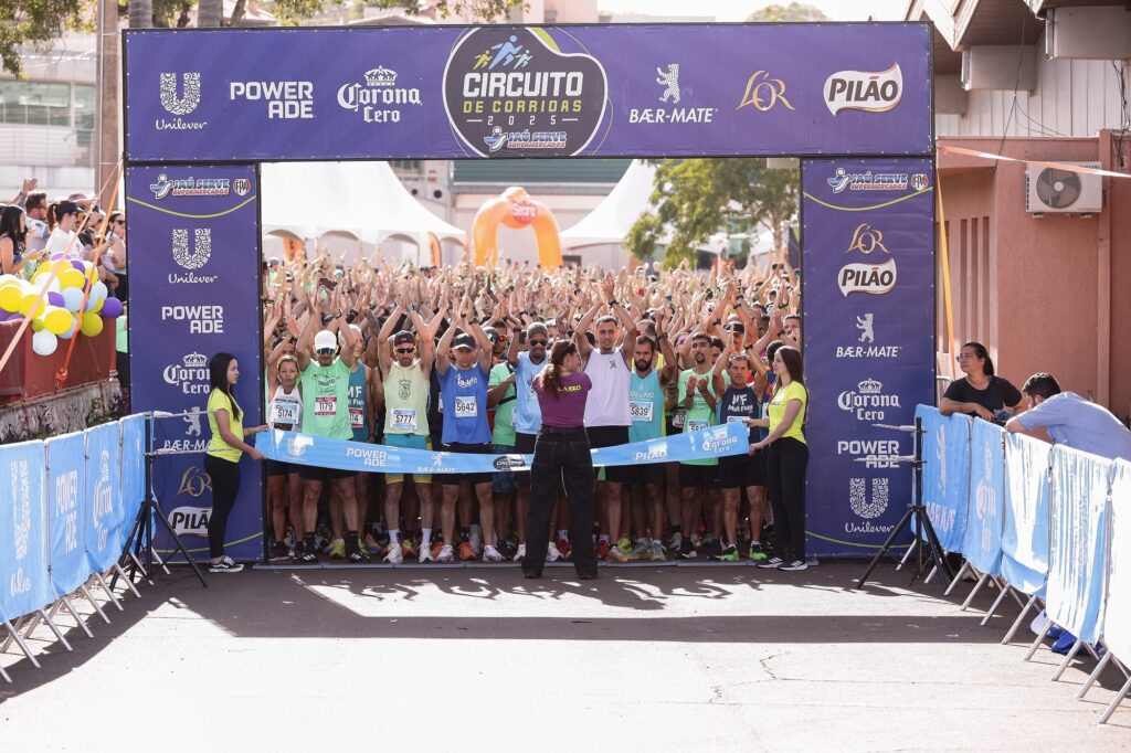 A imagem mostra o momento antes do início de uma corrida, parte do evento "Circuito de Corridas 2025 – Jaú Serve Supermercados". No centro, há uma faixa azul que marca a linha de largada, segurada por duas pessoas vestindo camisetas amarelas. Atrás da linha, um grande grupo de corredores está pronto para começar, muitos com os braços levantados, como se estivessem se alongando ou animados para a prova. Eles usam roupas esportivas variadas e números de identificação visíveis no peito. Acima deles, uma grande faixa roxa com o nome do evento e os logotipos dos patrocinadores (Unilever, Powerade, Corona Cero, Bær-Mate, L'OR e Pilão) está estendida. À frente dos corredores, uma mulher de camiseta roxa e calça preta parece estar coordenando ou dando instruções para a largada. O cenário é uma rua urbana, com espectadores atrás de uma grade decorada com balões coloridos à esquerda da imagem. Ao fundo, um arco inflável laranja marca parte do percurso da corrida. A iluminação é de um dia ensolarado, com sombras bem definidas no chão, transmitindo uma atmosfera de energia e expectativa para o início da prova.