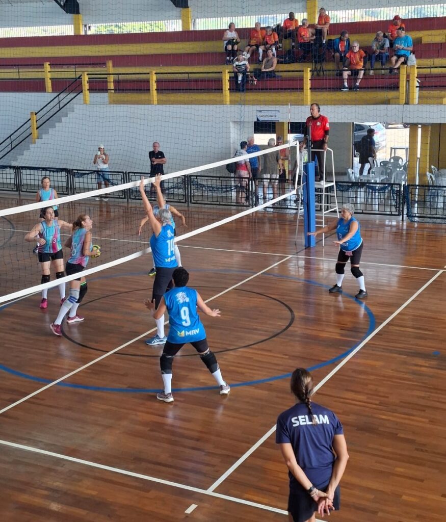 A imagem mostra uma partida de vôlei feminino sênior acontecendo em um ginásio coberto. A cena está focada em uma jogadora de camiseta azul número 14 que está saltando próximo à rede para bloquear a bola, enquanto uma jogadora adversária, vestindo uma camiseta azul clara com detalhes em rosa, está tentando atacar. Outras jogadoras da equipe de azul estão posicionadas na quadra, prontas para a defesa. Um árbitro com camiseta vermelha está em um dos lados da rede, em uma plataforma elevada, observando o jogo. Ao fundo, há arquibancadas com algumas pessoas assistindo à partida e uma placa indicando "Lanchonete". Um homem de camiseta azul escura com a inscrição "SELAM" nas costas está em primeiro plano, provavelmente um treinador ou membro da equipe técnica. O ambiente é iluminado por luzes internas, e o piso da quadra é de madeira com marcações coloridas. A imagem transmite a dinâmica e a intensidade de uma partida esportiva amadora ou comunitária.