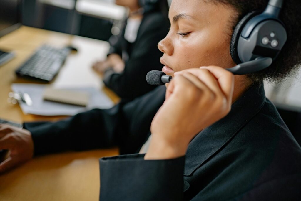 A imagem mostra uma mulher jovem, negra, em um ambiente de call center. Ela está vestindo um blazer preto e usa um fone de ouvido grande com microfone, que está ajustando com a mão direita. A mulher está de perfil, olhando para baixo, com uma expressão séria e concentrada, provavelmente focada em uma tela à sua frente. O fundo está desfocado, mas é possível ver outra pessoa também vestida de forma profissional, sugerindo um ambiente de trabalho compartilhado. A iluminação é suave, destacando a textura da pele da mulher e o tecido escuro da roupa. Há uma mesa de madeira clara com um teclado e alguns papéis, indicando um espaço de escritório. A imagem transmite uma atmosfera profissional e dedicada.