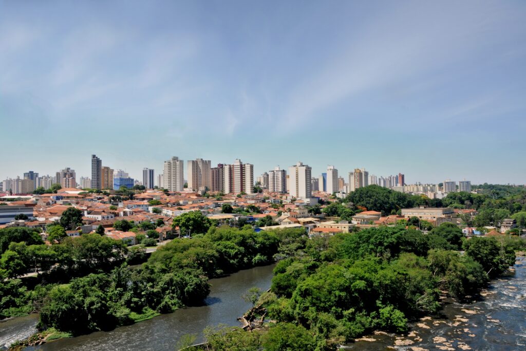 A imagem mostra uma vista panorâmica de uma cidade com uma transição clara entre áreas naturais e urbanas. Em primeiro plano, há um rio de águas calmas, margeado por uma vegetação verde e densa. À medida que avançamos para o meio da imagem, observa-se uma área residencial com várias casas de telhados vermelhos, típicos do estilo arquitetônico brasileiro. Ao fundo, destaca-se um conjunto de edifícios altos e modernos, formando o horizonte da cidade. O céu está claro e azul, com poucas nuvens, indicando um dia ensolarado. A composição transmite a integração entre a natureza e o ambiente urbano, mostrando o crescimento da cidade em meio a áreas verdes.