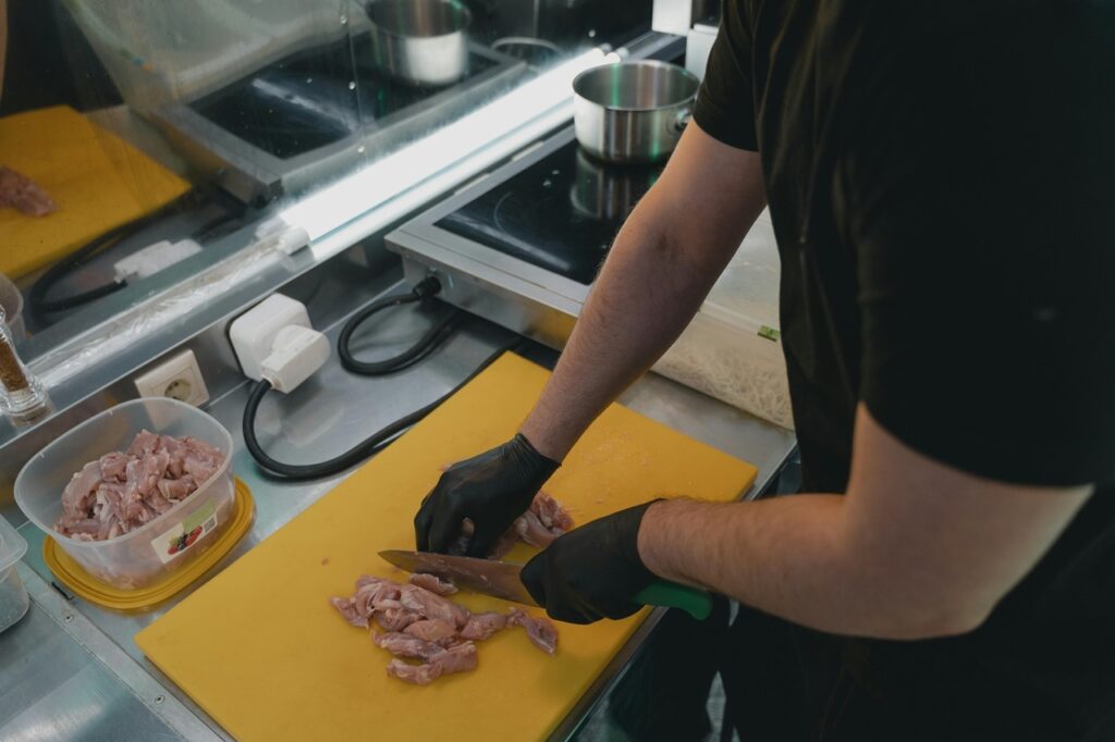 A imagem mostra uma cena em uma cozinha profissional, onde uma pessoa está cortando pedaços de carne crua (provavelmente frango) sobre uma tábua de corte amarela. A pessoa veste uma camiseta preta e luvas pretas de proteção, indicando cuidados com higiene e segurança alimentar. A tábua amarela é típica para manipulação de aves, seguindo normas de segurança na cozinha. Ao redor, há utensílios e equipamentos comuns em cozinhas industriais: uma frigideira na chapa de indução, um recipiente plástico transparente contendo mais carne crua, e um moedor de pimenta ou sal. O ambiente é iluminado com luz fria, e as superfícies são majoritariamente de aço inox, reforçando a ideia de um espaço profissional e limpo. A foto foi tirada de cima, focando especialmente nas mãos do cozinheiro e na tábua de corte, destacando o processo de preparação dos alimentos.