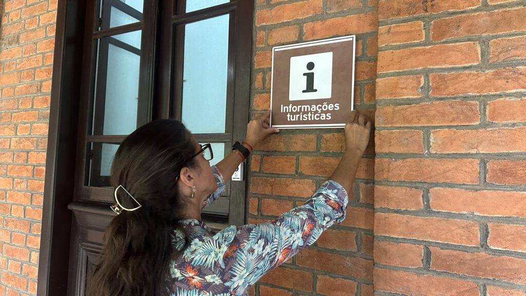 A imagem mostra uma mulher de perfil, com cabelos escuros presos por um grampo dourado, usando óculos e uma blusa estampada com flores em tons de azul, verde e laranja. Ela está fixando uma placa quadrada em uma parede de tijolos vermelhos. A placa é marrom com bordas brancas e tem um grande "i" minúsculo dentro de um quadrado branco na parte superior, indicando informações, e abaixo está escrito "Informações turísticas" em letras brancas. Ao lado esquerdo da mulher, há uma porta ou janela com moldura de madeira escura e vidro. A cena parece estar ao ar livre, em um ambiente iluminado pela luz do dia. A mulher está concentrada em posicionar a placa corretamente na parede.