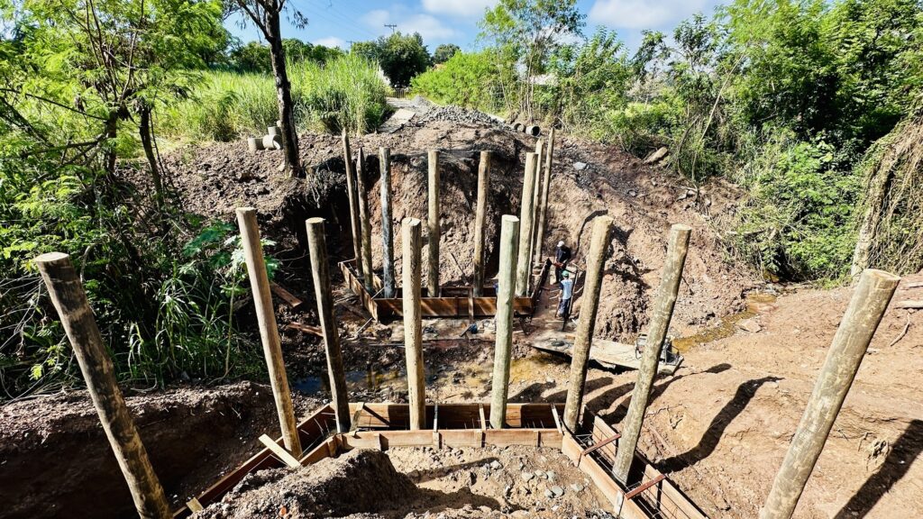A imagem mostra uma obra de construção em uma área rural, provavelmente a fundação de uma ponte ou de um muro de contenção. No centro da cena, há várias estacas de madeira fincadas verticalmente no solo, organizadas em duas fileiras que acompanham as margens de um pequeno córrego ou área alagada. No fundo, é possível ver vegetação densa com árvores e gramados verdes, indicando um ambiente natural e possivelmente tropical ou subtropical. Dois trabalhadores estão presentes no local, um deles usando capacete branco e roupa azul, aparentemente manuseando equipamentos ou materiais da obra. O solo ao redor das estacas está escavado, e há uma estrutura de madeira com vergalhões de ferro, sugerindo que está sendo preparada para receber concreto. A iluminação é forte, típica de um dia ensolarado, criando sombras marcantes e evidenciando os detalhes da construção. A cena transmite a ideia de um projeto de infraestrutura em andamento, situado em uma região afastada, cercada pela natureza.