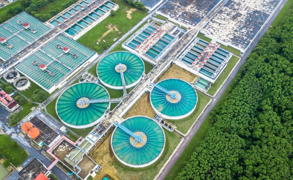 A imagem mostra uma vista aérea de uma grande estação de tratamento de água ou esgoto. No centro da foto, há quatro grandes tanques circulares, provavelmente usados para a decantação ou sedimentação, com água de cor verde-azulada. Cada tanque possui uma estrutura radial que parece ser um braço mecânico para limpeza ou movimentação da água. Ao redor desses tanques circulares, há vários tanques retangulares organizados em um padrão geométrico, também contendo água da mesma cor. Alguns desses tanques têm pequenas construções com telhados vermelhos, que provavelmente abrigam equipamentos ou controles. No canto inferior esquerdo, há duas grandes estruturas esféricas de cor laranja, que podem ser tanques de armazenamento ou unidades de tratamento específicas. A estação está cercada por áreas verdes bem cuidadas e, à direita da imagem, há uma área com árvores densas, indicando que a instalação está localizada em uma região com vegetação ao redor. O ambiente está bem iluminado, sugerindo que a foto foi tirada durante o dia com céu claro. A composição geral transmite uma sensação de organização, tecnologia e eficiência no tratamento da água.