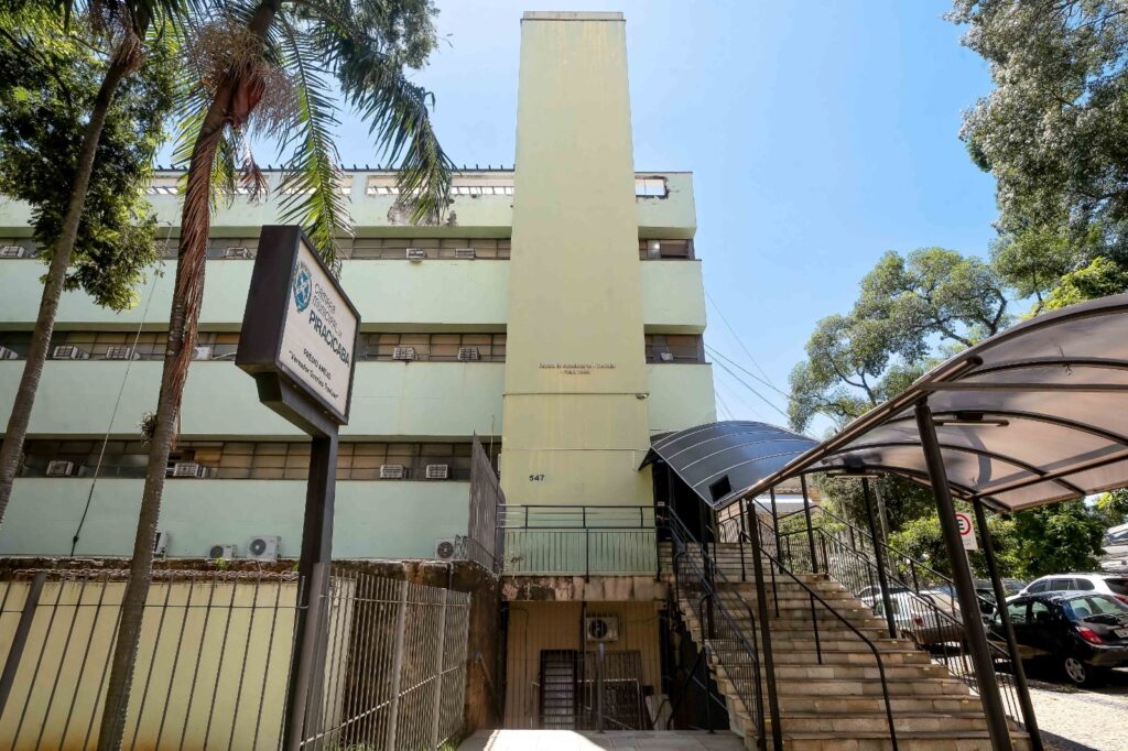 A imagem mostra o prédio anexo da Câmara Municipal de Piracicaba, chamado "Vereador Guerino Trevisan". O edifício é de cor verde clara, com várias janelas distribuídas em vários andares, muitas delas com aparelhos de ar-condicionado instalados. No centro da fachada, há uma coluna vertical que destaca a arquitetura do prédio. Em frente ao edifício, há uma placa grande com o nome da câmara e do prédio anexo, visível para quem passa pela rua. Ao redor do prédio, há árvores que emolduram a cena, conferindo um toque de natureza ao ambiente urbano. A iluminação é intensa, indicando que a foto foi tirada durante o dia, com sol forte. Também é possível ver uma escadaria com corrimão e um toldo escuro na entrada, além de um muro baixo com grades metálicas. Ao fundo, carros estão estacionados sob a sombra das árvores. A imagem transmite a atmosfera de um local oficial e institucional, situado em uma área arborizada e bem cuidada.