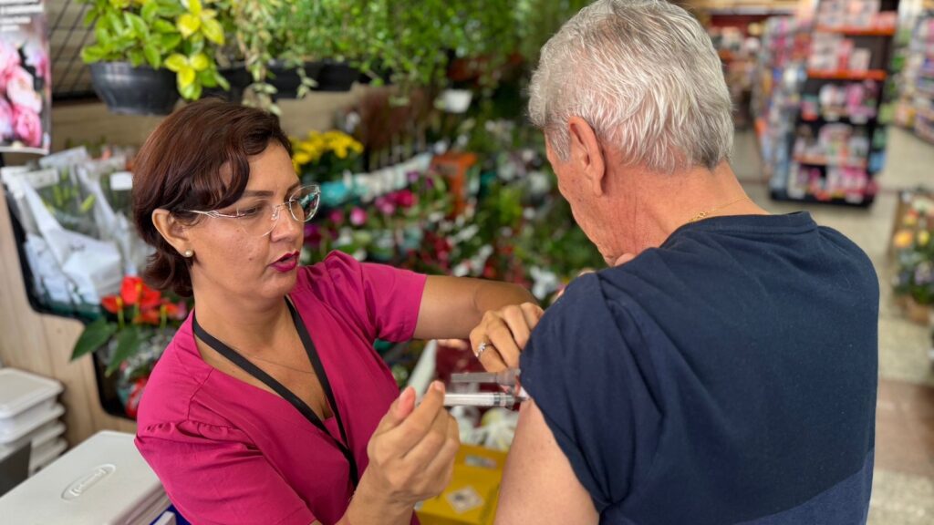 A imagem mostra uma mulher vestindo um uniforme rosa, possivelmente uma profissional de saúde, aplicando uma vacina no braço de um homem idoso. Ela está concentrada no procedimento, usando óculos e segurando a seringa com cuidado. O homem, de cabelos grisalhos, está de perfil e veste uma camiseta azul escura, aparentemente colaborando durante a aplicação. Ao fundo, há uma variedade de plantas e flores, sugerindo que a vacinação está ocorrendo em um ambiente que pode ser uma loja ou farmácia com seção de jardinagem. A iluminação é clara e o ambiente parece limpo e organizado. A cena transmite uma sensação de cuidado e profissionalismo no momento da vacinação.