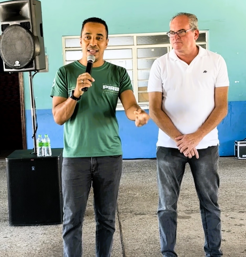 A imagem mostra dois homens em um ambiente interno, provavelmente uma sala ou auditório. O homem à esquerda está falando ao microfone, usando uma camiseta verde com o logo "PIRA" e calça jeans cinza escura. Ele parece estar explicando algo, com a mão esquerda levantada em um gesto. O homem à direita, mais velho, com cabelo grisalho e óculos, veste uma camisa polo branca e calça jeans cinza, e está observando atentamente o homem que fala, com as mãos juntas à frente do corpo. Ao lado deles, há um equipamento de som, incluindo caixas de som pretas empilhadas e dois copos de água sobre a caixa inferior. O fundo da cena é uma parede azul com uma janela de vidro com persianas horizontais. No canto da imagem, é possível ver parte de um banner ou cartaz com as palavras "PIRA", "SECRETARIA", "EDUCAÇÃO" e "PIRACICABA", indicando que o local pode estar relacionado à Secretaria de Educação da cidade de Piracicaba. A iluminação é clara e uniforme, típica de ambiente interno.