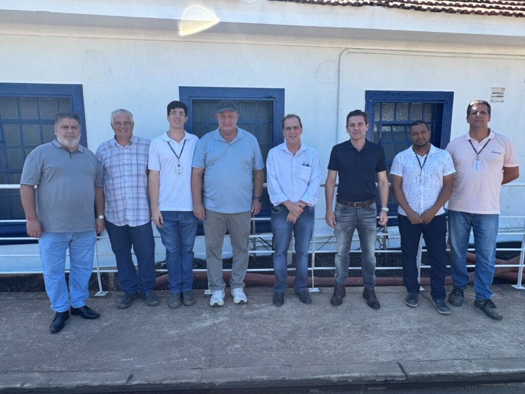 A imagem mostra um grupo de oito homens alinhados lado a lado, posando para a foto em frente a um prédio branco com janelas de moldura azul. Eles estão em uma calçada de concreto, com uma cerca branca atrás deles. Os homens vestem roupas casuais, principalmente camisas pólo e calças jeans ou sociais, e alguns usam crachás pendurados no pescoço. O ambiente parece ser externo, durante o dia, com luz natural brilhante iluminando a cena. À direita, na parede do prédio, há uma pequena placa com o nome "NOVELLO ALARMES". A atmosfera é descontraída, e todos os homens olham diretamente para a câmera.