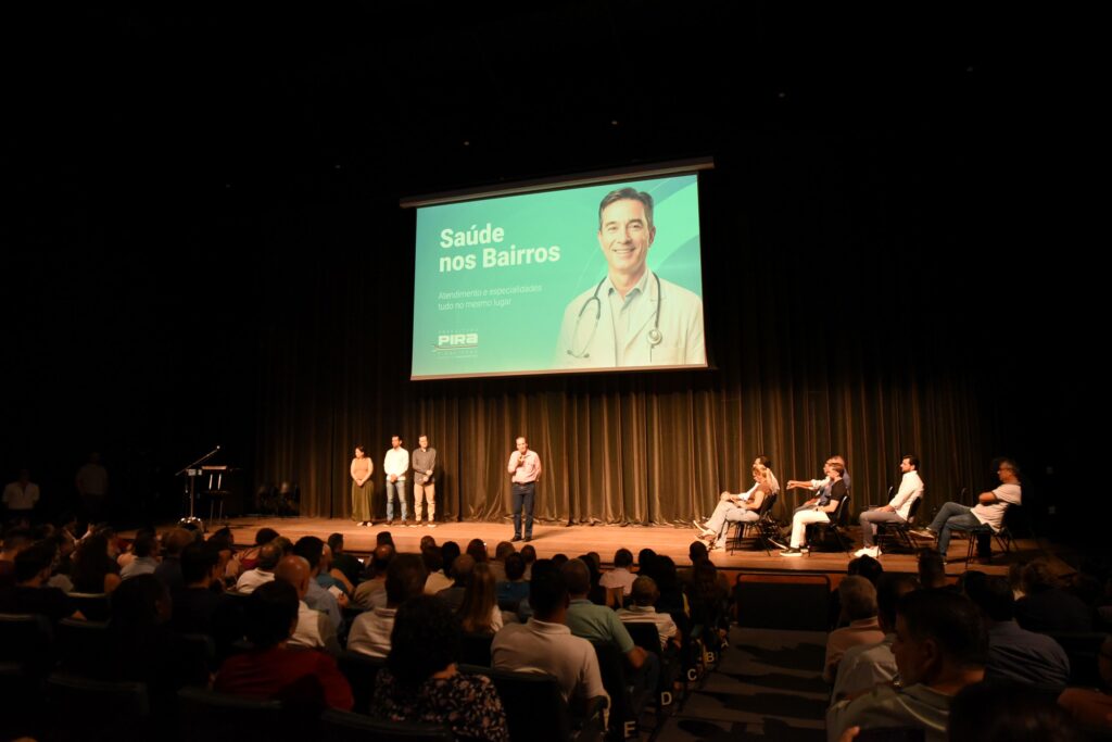 No palco, ao fundo, há uma grande tela exibindo o título "Saúde nos Bairros", com a foto de um médico sorridente e o logo da Prefeitura de Piracicaba. A tela tem um fundo verde claro com texto em branco. Há cortinas marrons escuras cobrindo o fundo do palco. No centro do palco, à frente da tela, há um pódio com um microfone. Quatro pessoas estão em pé no palco, na frente de uma fileira de cadeiras. Da esquerda para a direita, uma mulher com um colete laranja e saia longa verde; um homem de camisa branca e jeans; um homem de camisa xadrez cinza e calças bege; e um homem de camisa rosa e jeans, segurando um microfone e falando. Mais à direita no palco, há algumas pessoas sentadas em cadeiras, parecendo ser parte de um painel de discussão. Duas mulheres e três homens estão visíveis nesta área. No primeiro plano e ocupando a maior parte da imagem, há uma plateia sentada em cadeiras, voltada para o palco. A iluminação é baixa, focada no palco, o que faz com que a plateia esteja predominantemente na sombra, com os rostos e partes das roupas iluminados por cima. As cadeiras estão dispostas em fileiras, e algumas têm letras indicando a seção (B, C, D, E). As pessoas na plateia parecem estar atentas à apresentação, com algumas conversando discretamente. A atmosfera é de um evento formal ou informativo, com um tom de seriedade e interesse. As cores predominantes na plateia são tons escuros de roupas e do ambiente, contrastando com a luz clara do palco.