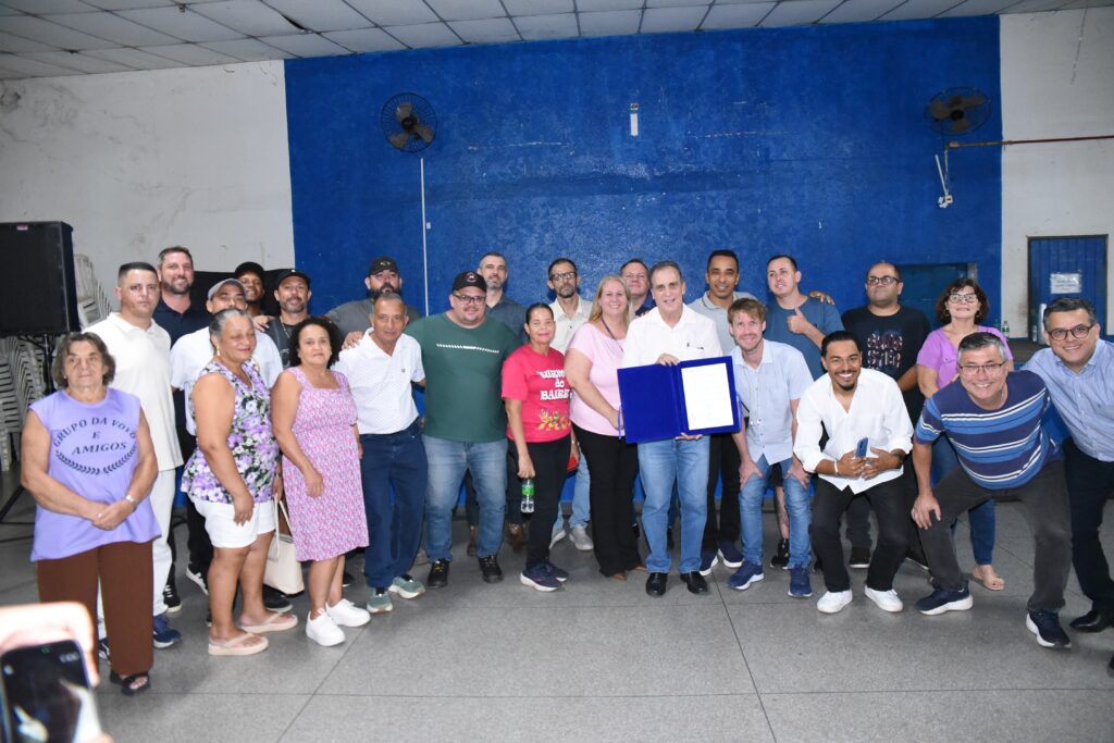 A imagem mostra um grupo de cerca de 25 pessoas reunidas em um ambiente interno, posando para uma foto em grupo. No centro, um homem de camiseta branca e calça jeans segura uma pasta azul com um documento visível, sugerindo que pode ser um momento de entrega ou apresentação formal. As pessoas ao redor estão sorrindo, transmitindo um clima de alegria e confraternização. O grupo é diversificado em termos de idade e gênero, com roupas casuais, incluindo camisetas, blusas e vestidos leves, adequados para um clima quente. Ao fundo, uma parede grande e azul serve como cenário, com dois ventiladores elétricos fixados no alto e uma caixa de som preta à esquerda. No canto esquerdo da imagem, uma mulher usa uma camiseta roxa com a inscrição "GRUPO DA VOVÓ E AMIGOS", indicando que o grupo pode estar relacionado a uma comunidade ou associação local. O ambiente parece ser uma sala ampla, com piso claro e iluminação interna forte. O conjunto sugere uma reunião comunitária, evento social ou celebração.