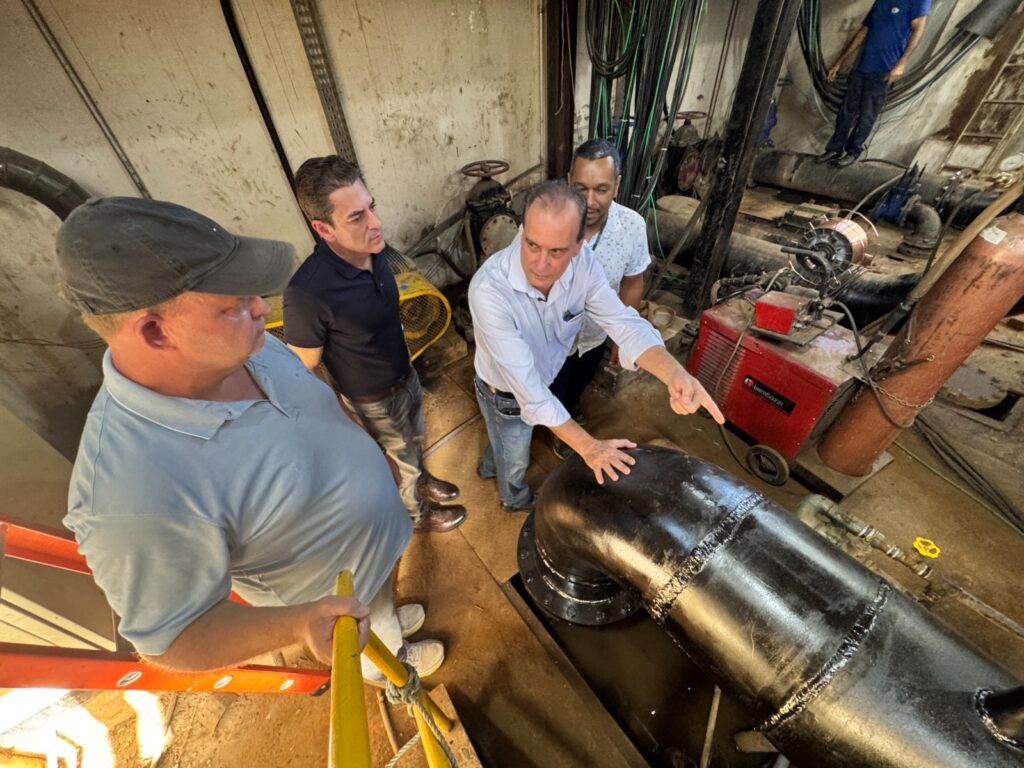 A imagem mostra quatro homens em um ambiente industrial, possivelmente uma estação de bombeamento ou uma área técnica subterrânea. Eles estão reunidos ao redor de uma tubulação grande e preta em forma de cotovelo. Um dos homens, vestido com uma camisa azul clara, está apontando para algum ponto da tubulação ou equipamento, enquanto os outros observam atentamente. O cenário apresenta paredes de concreto, fiação exposta e vários tubos e máquinas, incluindo um equipamento vermelho com a marca "bambozzi". A iluminação é mista, com luz artificial predominante e alguma luz natural entrando pela esquerda. O ambiente é claramente técnico e funcional, com uma atmosfera de inspeção ou manutenção em andamento.
