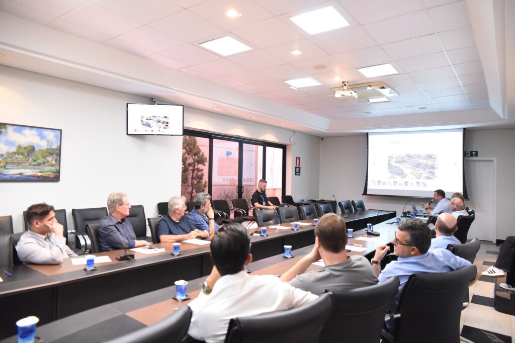 A imagem mostra uma reunião profissional em uma sala de conferências moderna. Há uma mesa grande em formato de "U", de madeira escura, ao redor da qual estão sentados cerca de dez homens, a maioria voltada para uma grande tela de projeção na parede ao fundo. Na tela, é exibida uma imagem aérea colorida, possivelmente de um projeto arquitetônico ou urbanístico. Também há um monitor menor na parede lateral esquerda mostrando uma imagem semelhante. Os participantes são homens de meia-idade a idosos, vestidos de forma casual, com camisas sociais e polos. Alguns conversam entre si, enquanto outros observam atentamente a apresentação. No fundo da sala, próximo a uma porta de vidro, um homem parece estar controlando os aspectos técnicos da reunião. A sala é bem iluminada por luzes embutidas no teto, que é branco e possui um design moderno. Na parede esquerda há uma pintura de paisagem, com um rio e árvores. A mesa está equipada com microfones e pequenos copos azuis de plástico. O piso tem um padrão quadriculado em preto e branco perto da porta à direita. O ambiente transmite uma atmosfera séria e focada, típica de uma reunião de trabalho ou apresentação importante.