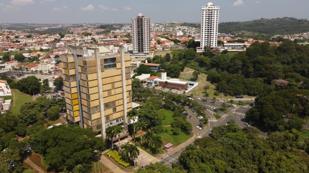 A imagem mostra uma vista aérea de uma área urbana, com destaque para um prédio grande e retangular de estilo brutalista, pintado em amarelo com pilares de concreto à mostra. Esse prédio está localizado em primeiro plano, à esquerda da imagem, e possui várias janelas alinhadas em fileiras. Ao redor dele há uma área de estacionamento com diversos carros estacionados. Mais ao fundo, há uma área verde com árvores, incluindo palmeiras, e um pequeno quiosque ou estrutura vermelha. Atrás dessa área verde, aparecem dois prédios altos e modernos, um branco e outro cinza, que se destacam na paisagem. No horizonte, percebe-se uma grande extensão de casas com telhados vermelhos, típica de bairros residenciais, e colinas ao fundo sob um céu claro e ensolarado. A imagem transmite uma sensação de organização urbana, com contraste entre o prédio amarelo robusto e os edifícios mais altos e modernos ao longe.