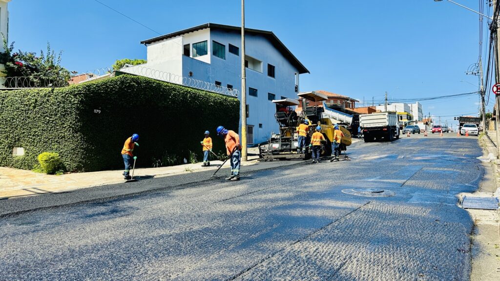 A imagem mostra uma equipe de trabalhadores realizando a pavimentação de uma avenida em um dia ensolarado. No primeiro plano, há uma faixa de asfalto recém-colocado, escuro e liso. No meio da imagem, seis operários vestem uniformes laranja fluorescente com capacetes azuis, alguns estão utilizando ferramentas para alisar o asfalto nas bordas. À direita, há uma máquina amarela para pavimentação e, atrás dela, um caminhão branco que provavelmente transporta o material para o trabalho. No fundo, há um prédio branco de vários andares e uma cerca viva com arame farpado no topo, além de alguns carros estacionados e postes de energia. O céu está azul e limpo, indicando um dia claro e ensolarado. A foto foi tirada de um ângulo baixo, dando destaque ao trabalho e ao equipamento.
