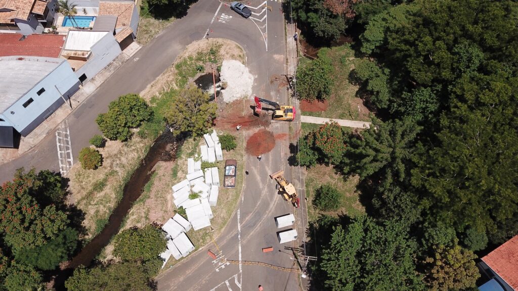 A imagem mostra uma obra em andamento em uma estrada localizada em uma área residencial com bastante vegetação ao redor. A estrada faz uma curva acentuada e está parcialmente bloqueada para o trabalho. No local, há máquinas pesadas, como uma escavadeira amarela e vermelha da marca Volvo, que está escavando o asfalto, e uma pá carregadeira amarela próxima. Ao lado da estrada, há uma pilha de grandes blocos de concreto brancos empilhados sobre a grama, junto a um pequeno córrego que corre paralelo à via. No chão da estrada está pintado o aviso "PARE", indicando que os veículos devem parar, provavelmente devido à obra. Também é possível ver alguns trabalhadores no local, alguns usando roupas de alta visibilidade e outros com roupas casuais. Há um caminhonete estacionada perto dos blocos de concreto e outro veículo prata mais adiante na estrada. Ao fundo, casas residenciais com telhados de cerâmica e até uma piscina são visíveis, cercadas por árvores e vegetação densa. A iluminação da imagem sugere que foi tirada durante o dia, com sol forte e sombras bem definidas. No geral, a imagem retrata uma manutenção ou construção em uma estrada de bairro, em uma área que combina elementos urbanos e naturais.