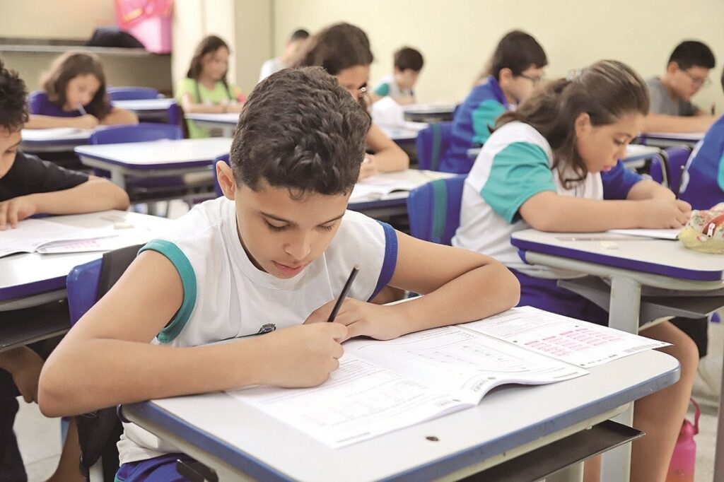 A imagem mostra uma sala de aula com várias crianças sentadas em carteiras, concentradas em suas atividades. Em primeiro plano, há um menino vestido com uma camiseta regata branca com detalhes em azul e verde, focado em escrever em seu caderno ou livro de exercícios. Ele está segurando um lápis e parece estar bastante atento ao que está fazendo. Ao seu lado, uma menina também está concentrada, vestindo uma camiseta branca com detalhes azuis. Ao fundo, outras crianças também estão sentadas em suas carteiras, algumas escrevendo, outras olhando para frente. A sala é bem iluminada, provavelmente com luz natural, e o ambiente transmite uma sensação de concentração e estudo sério. As carteiras são azuis e brancas, e no chão há uma garrafa de água rosa. A cena sugere uma aula ou prova em andamento, com os alunos focados em suas tarefas.