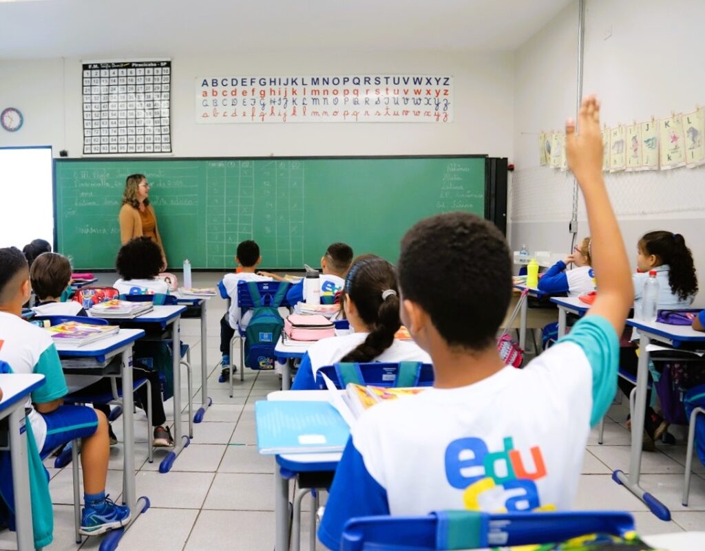 A imagem mostra uma sala de aula iluminada e organizada, provavelmente em uma escola primária no Brasil, dado o texto em português e a referência a "Piracicaba - SP". No centro da cena, uma professora está em pé ao lado do quadro verde, explicando uma lição. Ela usa óculos e um suéter marrom, e parece estar gesticulando para os alunos. Vários estudantes estão sentados em carteiras azuis e brancas, usando uniformes com a palavra "educa" e cores azul e verde. Um aluno em destaque, em primeiro plano, levanta a mão, indicando que quer responder ou fazer uma pergunta. A sala possui cartazes educacionais nas paredes, incluindo o alfabeto completo e uma tabela de números de 1 a 100, sugerindo um ambiente de ensino fundamental. No quadro, há anotações escritas à mão, possivelmente relacionadas a uma atividade de matemática ou português, com uma tabela e palavras em português. A atmosfera é de aprendizado ativo e participação, com os alunos atentos e a professora conduzindo a aula de forma envolvente.