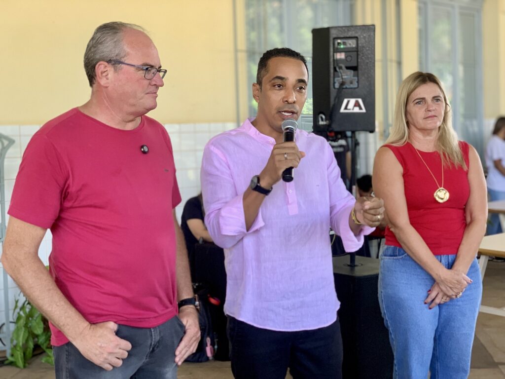 A imagem mostra três pessoas em pé, alinhadas lado a lado, aparentemente participando de um evento público ou reunião comunitária. A pessoa no centro, um homem de camisa rosa clara, está falando ao microfone, enquanto as outras duas, um homem à esquerda vestido com camiseta vermelha e calça jeans cinza, e uma mulher à direita usando uma blusa vermelha e calça jeans azul, observam. Ao fundo, há um alto-falante preto com o logotipo "LA". O ambiente é iluminado por luz natural, com paredes amarelas e detalhes em branco, sugerindo um espaço como uma escola, centro comunitário ou salão público. A atmosfera parece formal, porém descontraída, indicando uma apresentação ou discurso em um evento local.