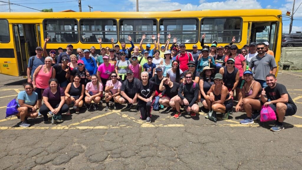 A imagem mostra um grande grupo de cerca de 50 pessoas reunidas em frente a um ônibus amarelo vibrante. Elas estão organizadas em três fileiras: algumas agachadas ou sentadas na frente, e as demais em duas fileiras atrás, muitas acenando ou fazendo gestos com as mãos. A maioria está vestida com roupas esportivas casuais, como camisetas, regatas, shorts, leggings, bonés e óculos de sol, sugerindo que participaram de uma atividade ao ar livre ou um evento esportivo. O cenário é um dia ensolarado, com céu azul e poucas nuvens, e o grupo está posando sobre um chão de asfalto escuro com algumas linhas amarelas pintadas. Ao fundo, além do ônibus, é possível ver uma parede de concreto, um carro escuro estacionado e algumas linhas de energia elétrica. A atmosfera geral é alegre e de confraternização.