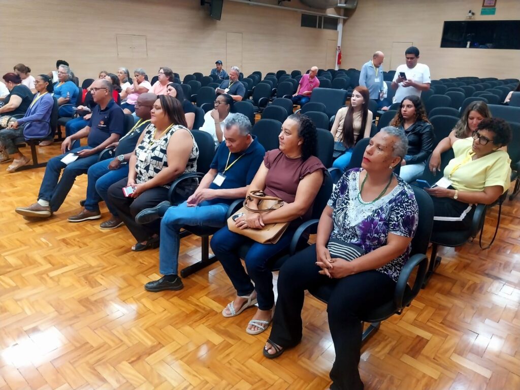 A imagem mostra um auditório com várias pessoas sentadas em fileiras de cadeiras pretas acolchoadas. O ambiente tem um piso de madeira com acabamento brilhante e paredes também de madeira, criando uma atmosfera acolhedora e elegante. A maioria das pessoas está voltada para a frente, provavelmente assistindo a uma apresentação ou palestra, enquanto algumas olham para seus celulares. As roupas dos presentes variam bastante, com cores como azul, amarelo, preto e estampas diversas, sugerindo um público diversificado. No fundo, há uma parede escura que pode ser uma tela ou uma porta. A iluminação é quente, destacando o ambiente e os rostos das pessoas. O cenário transmite a ideia de um evento formal ou educativo.